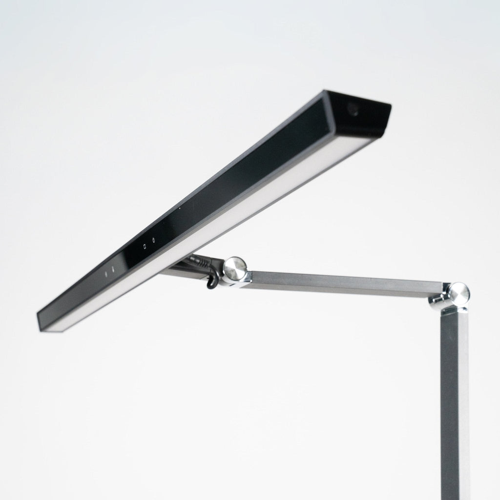 Zbliżenie na nowoczesną minimalistyczną lampę biurkową Heilys z pełnospektralnym światłem, prostokątną płaską czarną głowicą i metalowym regulowanym ramieniem - idealną do oświetlenia biurowego - ustawioną na prostym białym tle.