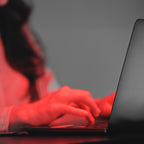 Osoba pisząca na laptopie z obiema rękami pod heilys pełnospektralne światło. Obraz ma czerwony odcień, pokazujący tylko dłonie, ramiona i laptop na rozmytym tle.