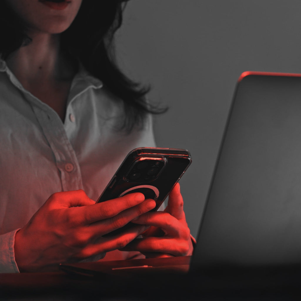 Osoba w białej koszuli korzysta ze smartfona przy laptopie, z heilys pełnospektralne światło i czerwone oświetlenie biurowe oświetlające ręce i część twarzy.