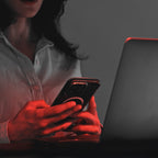 Osoba w białej koszuli korzysta ze smartfona przy laptopie, z heilys pełnospektralne światło i czerwone oświetlenie biurowe oświetlające ręce i część twarzy.