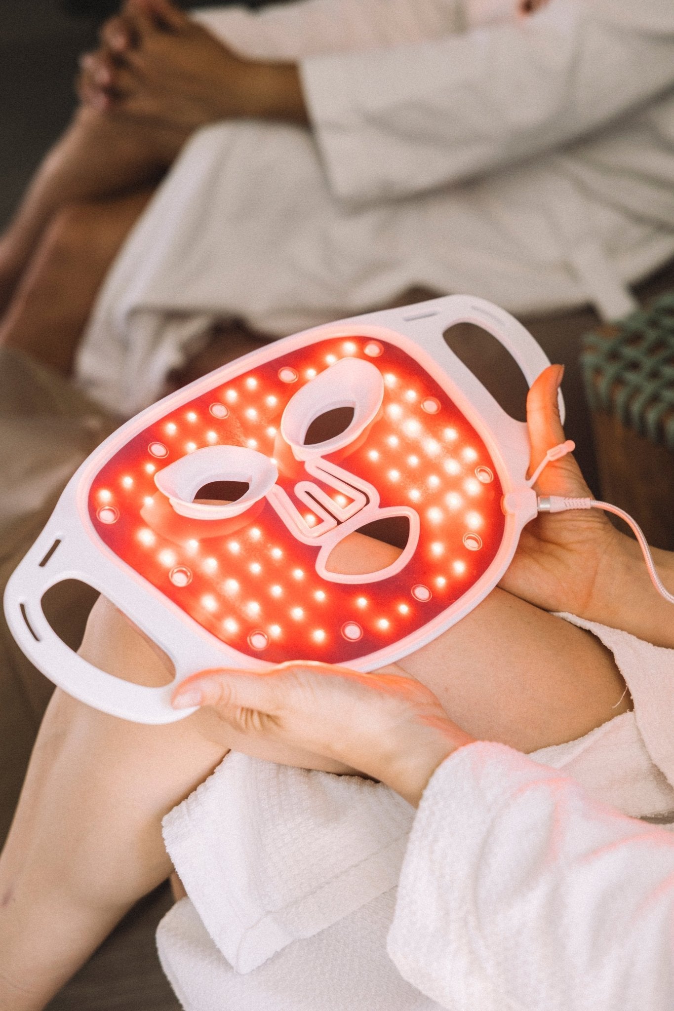Osoba w białej szacie trzyma podświetlaną na czerwono maskę LED do terapii światłem heilys, prezentującą nowoczesną technologię przeciwzmarszczkowej pielęgnacji skóry; w tle siedzi inna ubrana osoba ze złożonymi rękami.