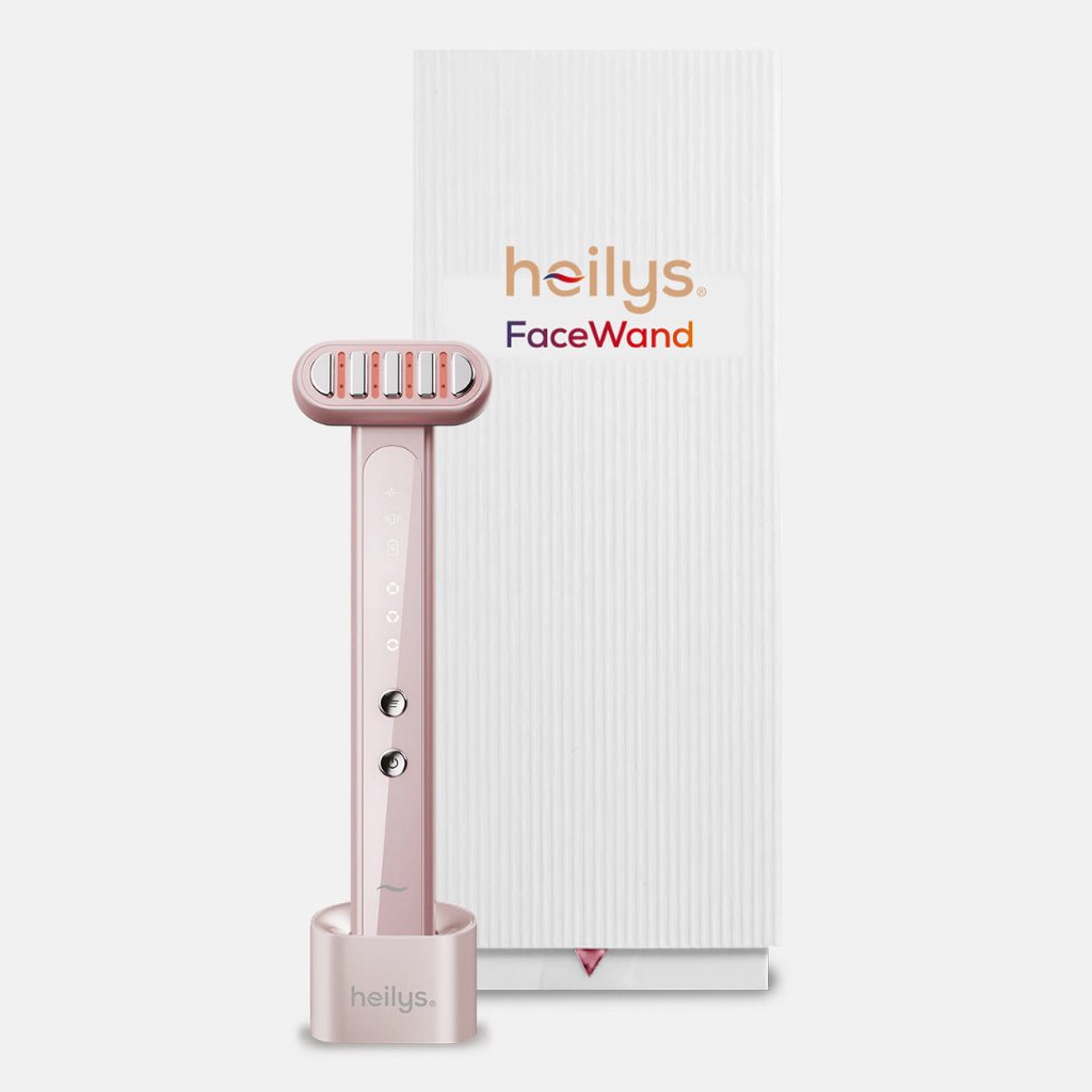 Różowe urządzenie do twarzy heilys FaceWand 5 w 1 stoi pionowo w bazie ładującej obok białego, pionowego pudełka z logo heilys FaceWand.