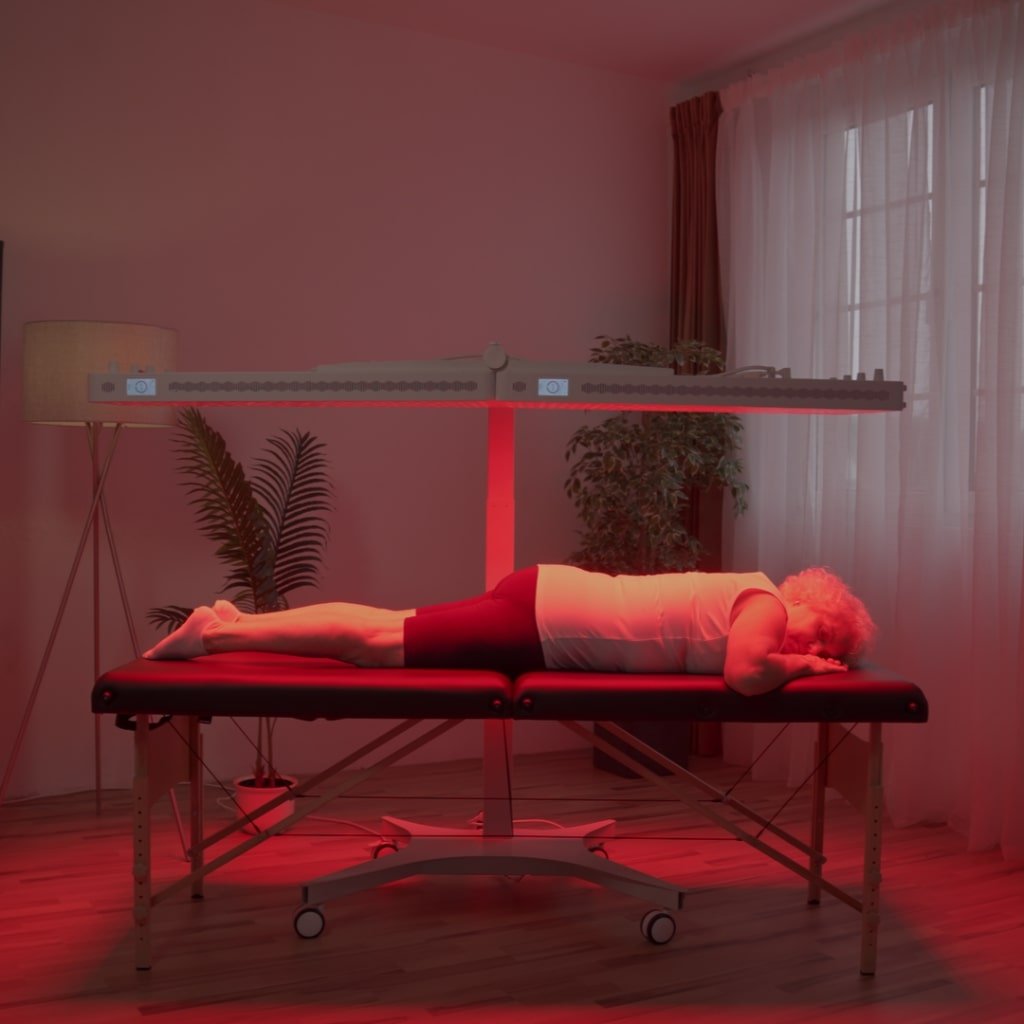 Starsza kobieta leży twarzą w dół na stole do masażu pod lampą heilys LED czerwone i NIR zestaw do naświetlania w delikatnie oświetlonym pokoju z drewnianymi podłogami, roślinami domowymi, lampą podłogową i zasłonami nad dużymi oknami.