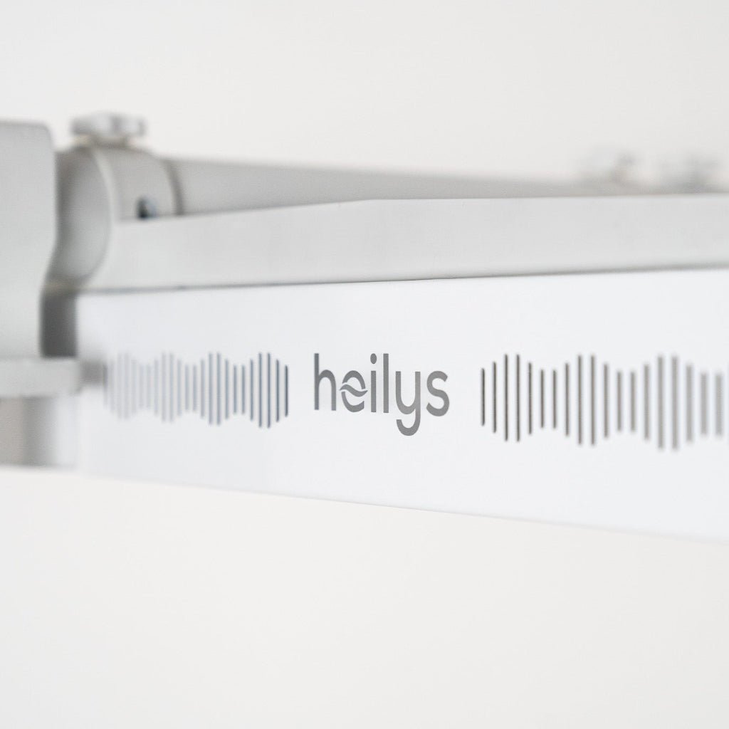 Zbliżenie na białą metalową powierzchnię z napisem "heilys" w szarych małych literach pomiędzy wzorami fal dźwiękowych, prezentującą lampę LED czerwone i NIR zestaw do naświetlania przez heilys, ustawioną na rozmytym tle.