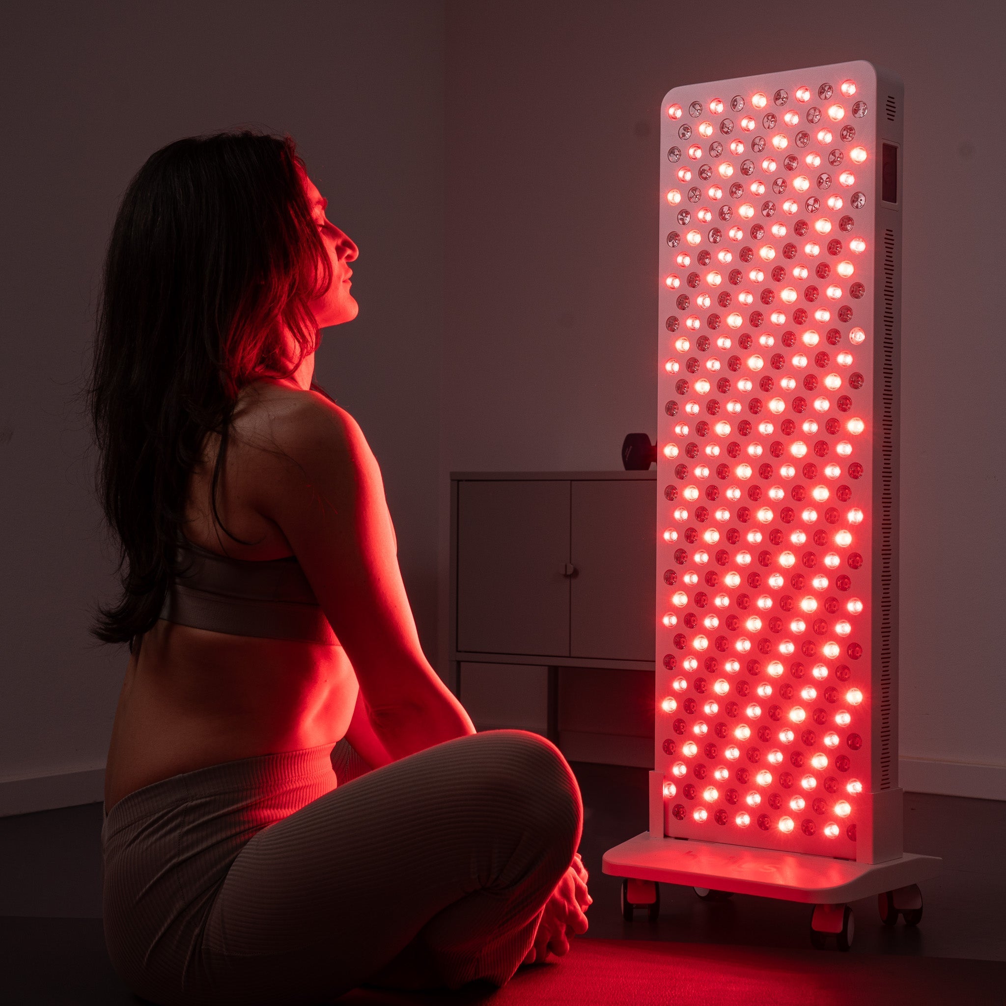 Kobieta w stroju sportowym siedzi ze skrzyżowanymi nogami na podłodze, naprzeciwko wysokiego panelu heilys ze świecącymi czerwonymi światłami, używając urządzenia do terapii czerwonym światłem heilys w słabo oświetlonym pomieszczeniu.