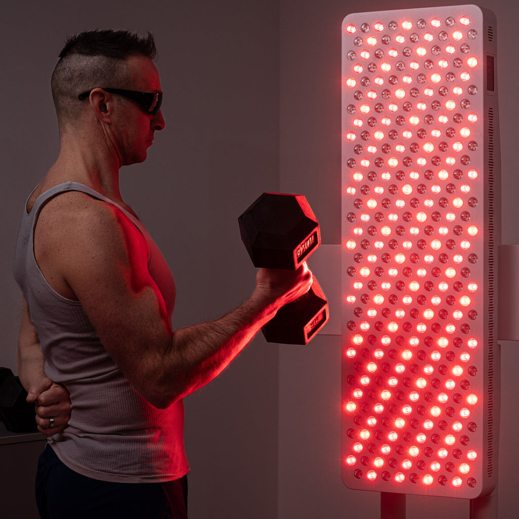Mężczyzna w okularach przeciwsłonecznych i podkoszulku podnosi hantle obok dużego pionowego panelu LED heilys do terapii światłem, jego czerwone diody LED są jasno oświetlone.