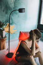 Kobieta w czerni siedzi na krześle w pomieszczeniu, dotykając włosów. Za nią znajduje się roślina doniczkowa, panel heilys światła czerwonego o długości fali 670 nm i tealowa ściana ozdobiona złotem.