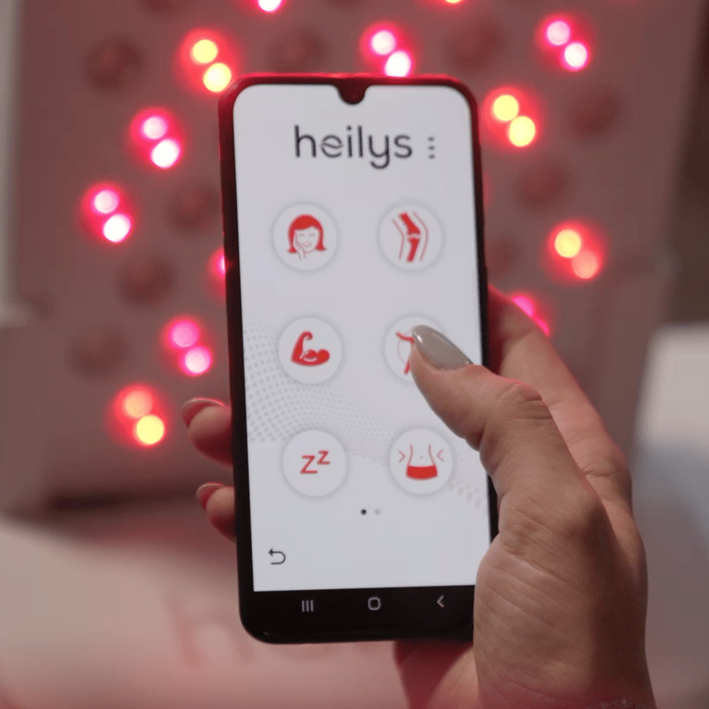 Dłoń trzyma smartfon pokazujący aplikację heilys z ikonami dobrego samopoczucia - twarzy, pleców, ramion, snu i relaksu. Rozmyte czerwone światło NIR w tle sugeruje użycie kompaktowego urządzenia do światłoterapii heilys.